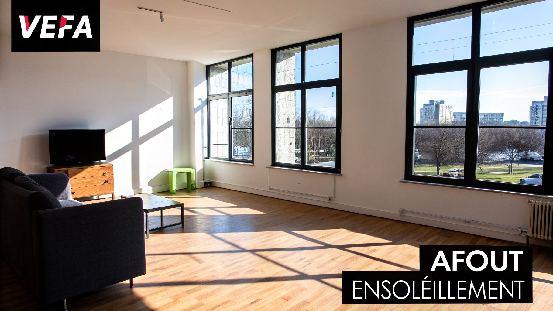 Intérieur moderne et ensoleillé avec canapé gris, télévision, et plancher en bois, baigné de lumière naturelle.